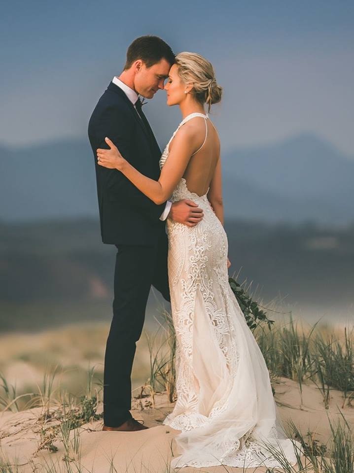 A man in a formal suit and a woman in a long wedding dress embracing on a beach.