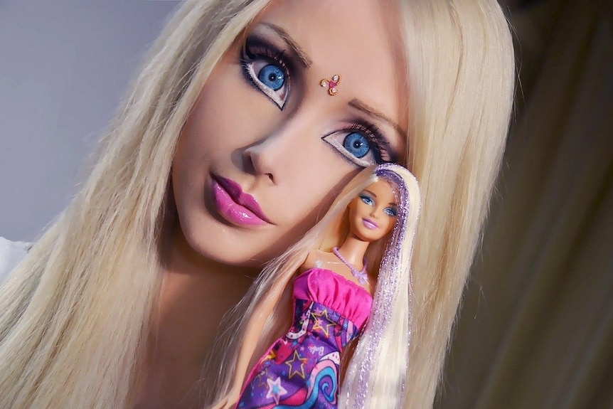 A woman who looks like Barbie, holding a Barbie doll
