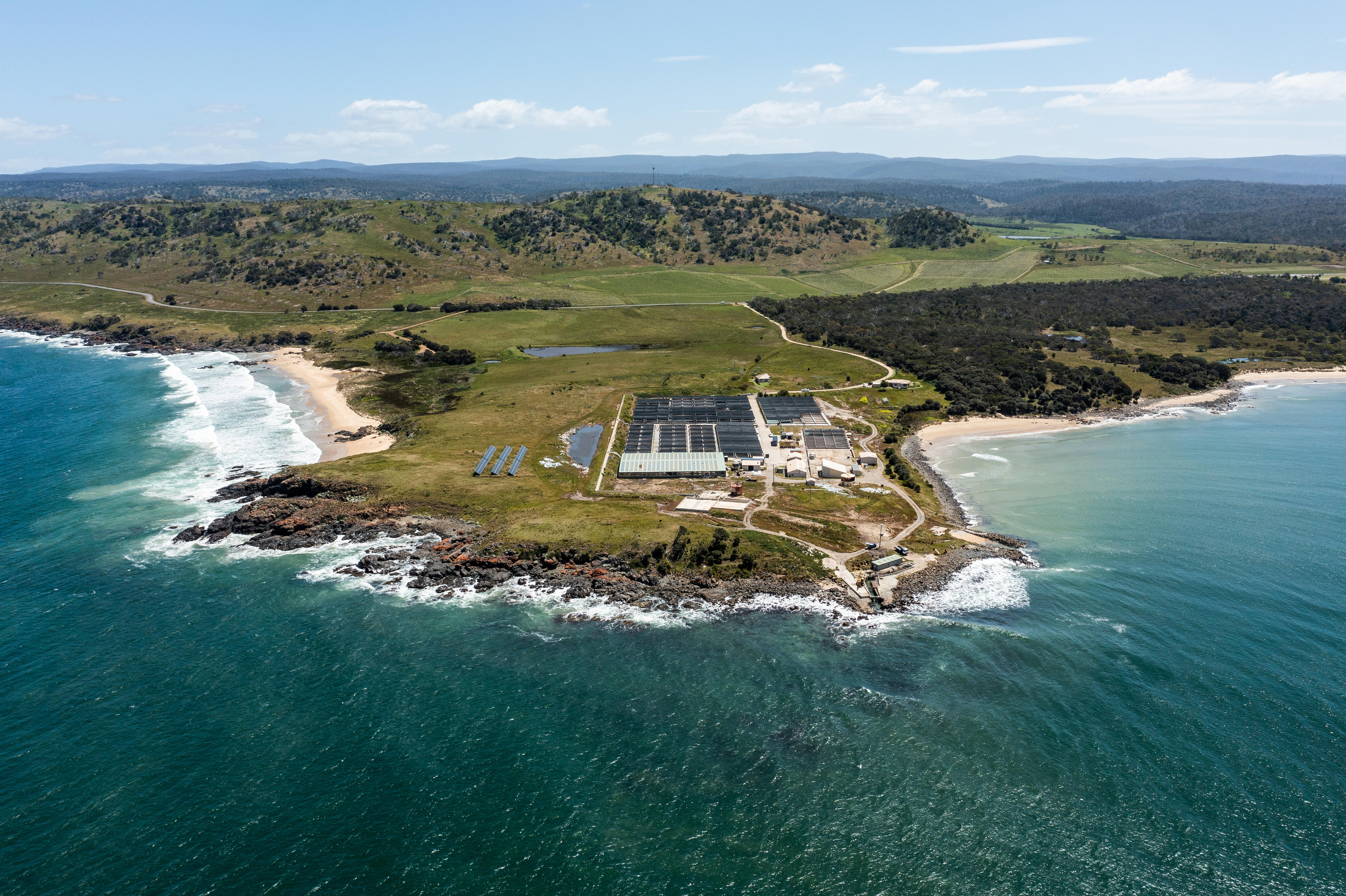 An aerial view of a cliffside research facility. It's a clear sky day and ocean laps the edges of the cliff 
