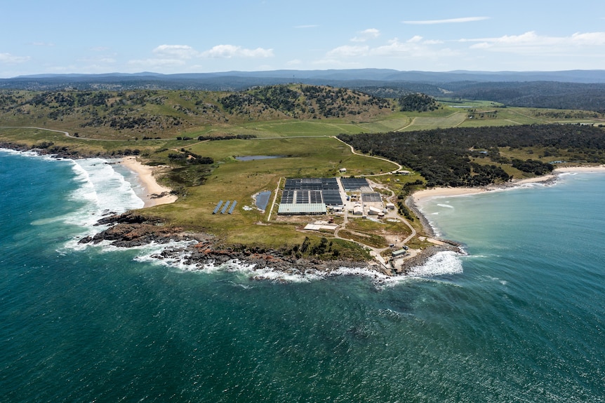 An aerial view of a cliffside research facility. It's a clear sky day and ocean laps the edges of the cliff 