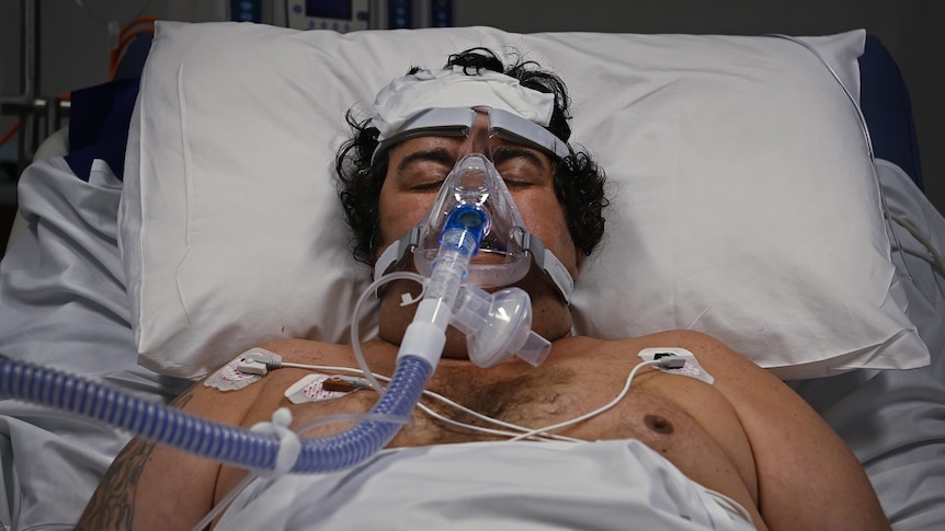 A man in bed with his eyes closed, with an oxygen mask on.