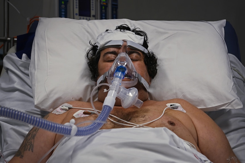 A man in bed with his eyes closed, with an oxygen mask on.