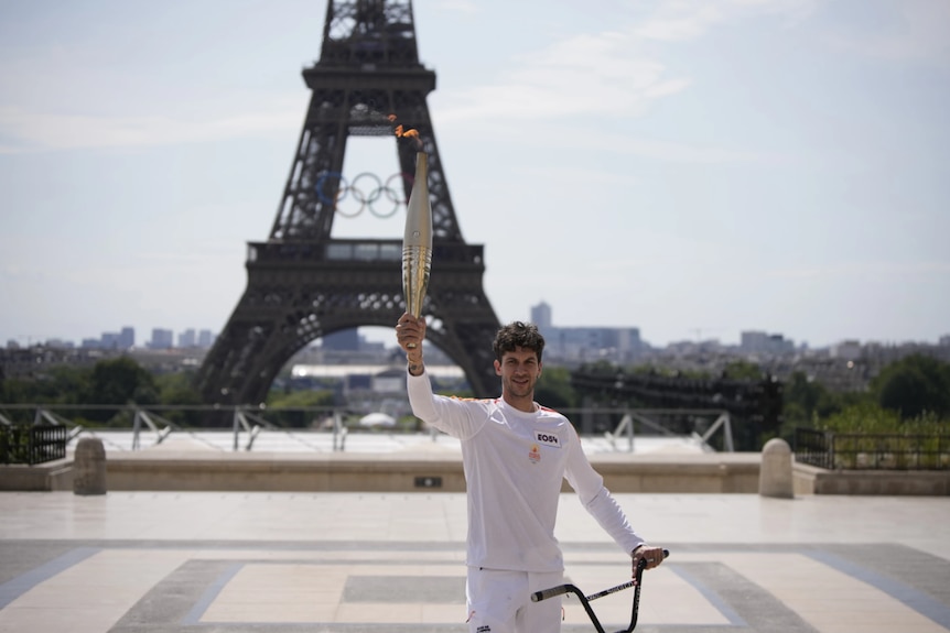 A man dressed in white smiles and holds a large torch in front of the Eiffel Tower