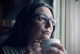A woman with long dark hair and glasses stares out the window while grasping a cup of tea.