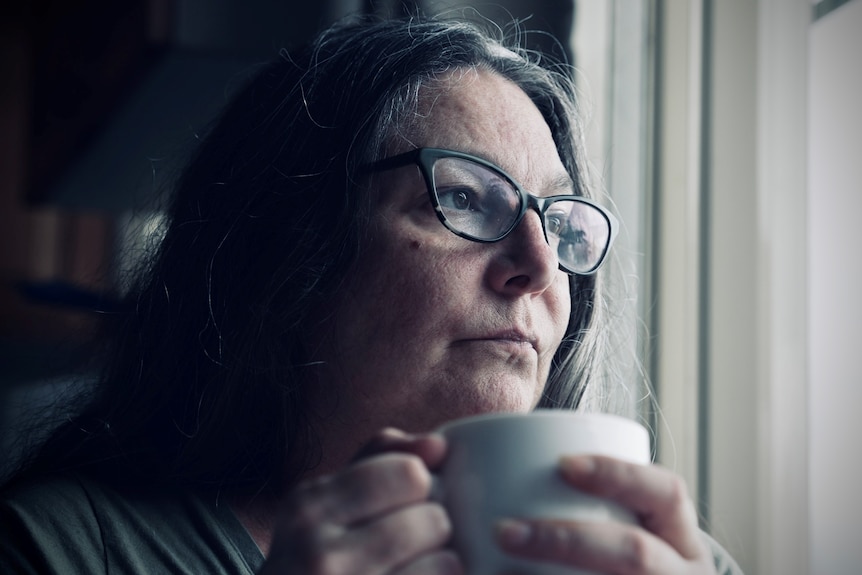A woman with long dark hair and glasses stares out the window while grasping a cup of tea.