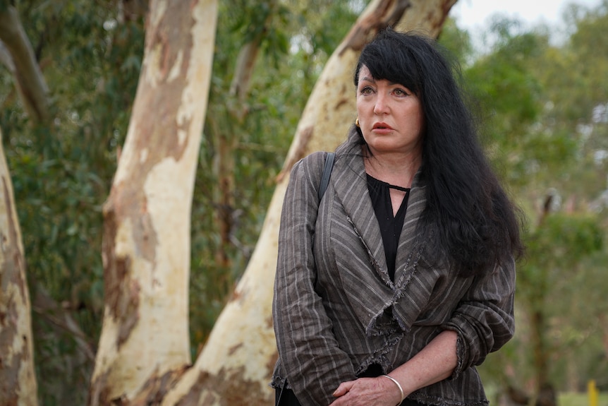 A woman with long, dark hair, stands in front of a large tree and woodland beyond.