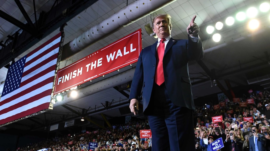 From a low angle, a photo shows Donald Trump pointing with his left index finger with a red 'Finish the Wall' banne behind him.