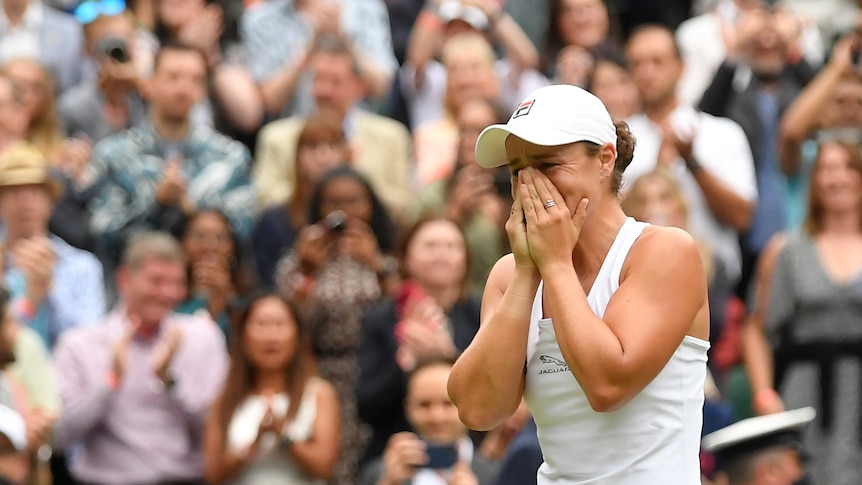 A young woman in tennis whites reacts emotionally on a grass court in front of cheering fans.