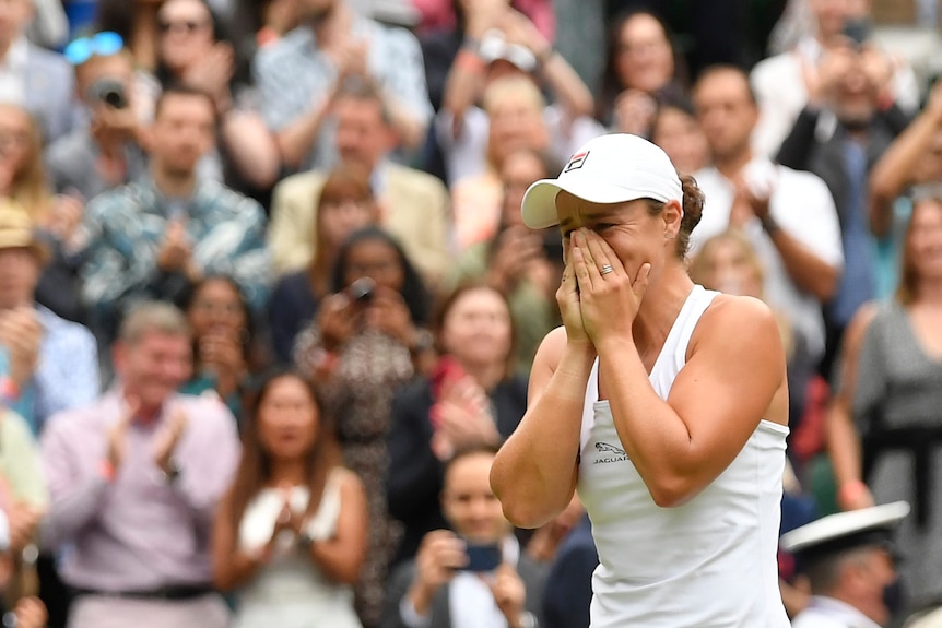 Une jeune femme en blanc de tennis réagit avec émotion sur un terrain en gazon devant des supporters en liesse.