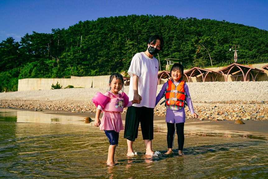 A Korean woman walks on a shoreline, holding hands with two little girls 