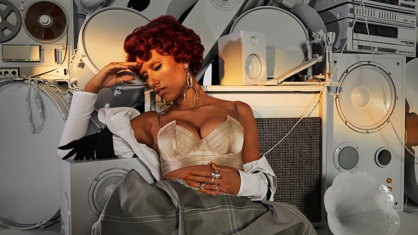 RAYE wears a gold bra, grey skirt and curled red hair as she sits in front of white speakers and recording hardware.