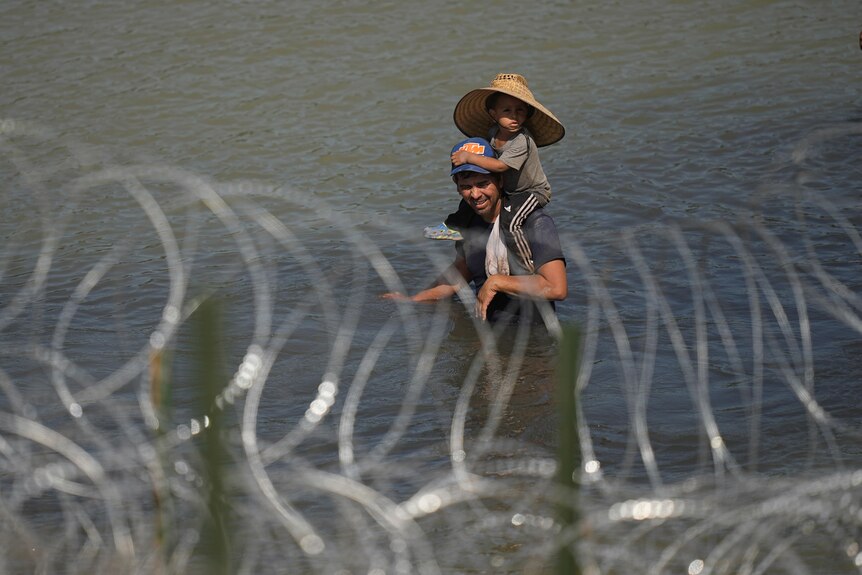 A man stands in water with a child sitting on his shoulders. They are behind a wire fence.