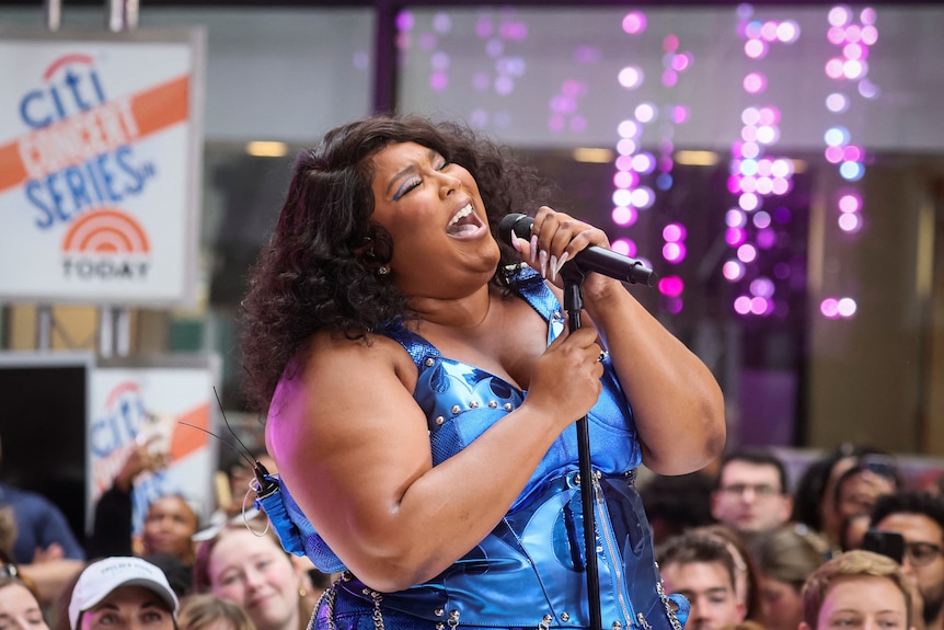 lizzo sings into a microphone on a stand on a raised stage surrounded by people wearing a shiny blue costume