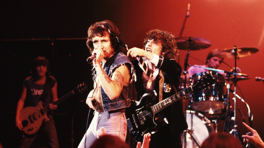 Bon Scott and Angus Young on stage peforming under red lighting setting. Angus puts his necktie around Bon's neck joking fashion