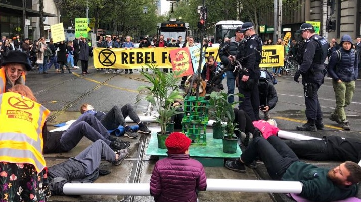 A group of protesters in jackets lie in a circle in the middle of an intersection with arms connected as police look on.