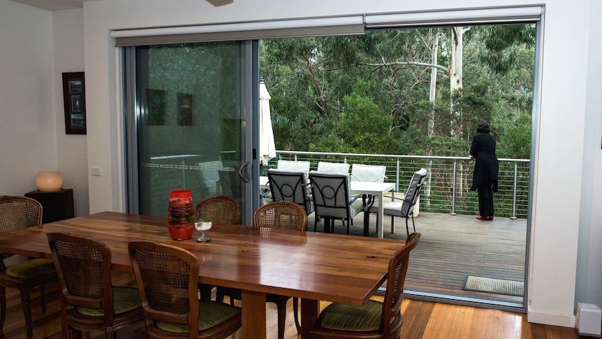A woman stands on a deck, overlooking Australian forest, room with timber features in foreground.