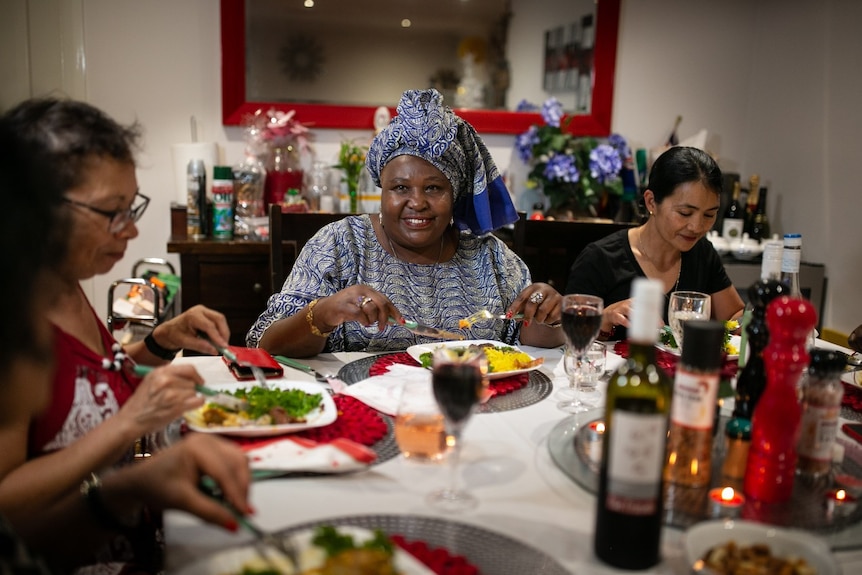 A woman in a blue headdress and matching top sits poised to eat at a dinner table with friends.