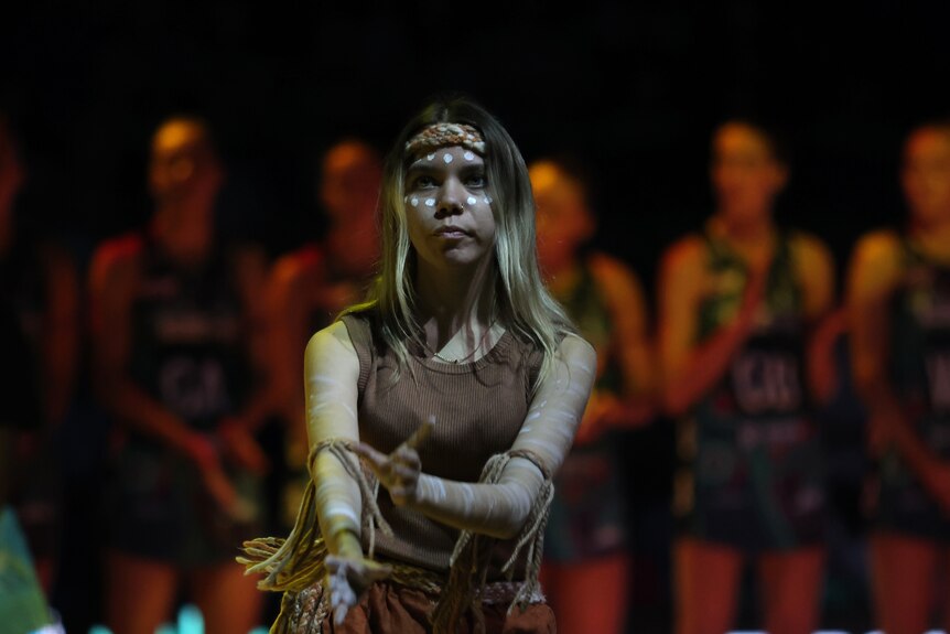 A girl wears traditional First Nations clothing and face paint as a netball team stands behind her 