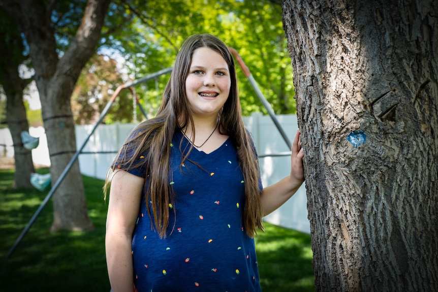 A young girl with dark hair flowing over her shoulders poses next to a tree