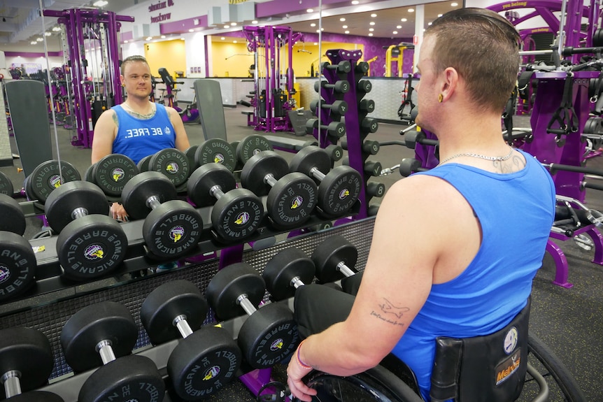 A man in a wheelchair stares into a mirror in front of a weights rack at the gym 