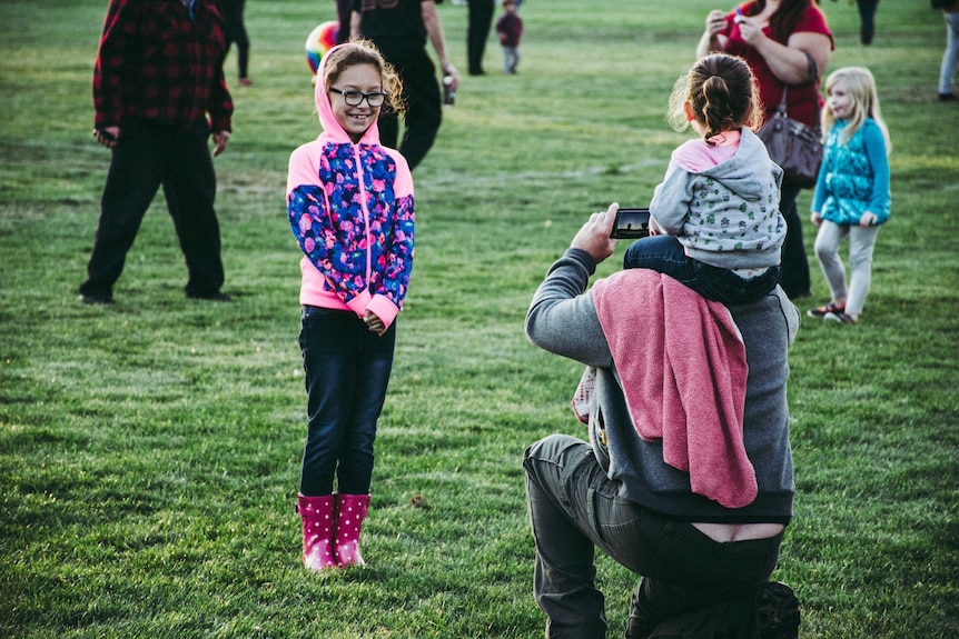 A father kneeling with a child on his shoulders as he takes a photo of a smiling young girl