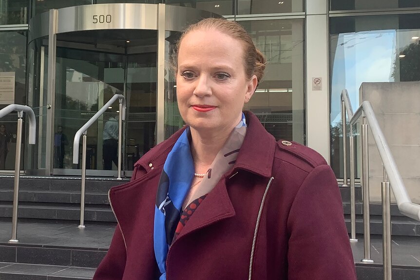 A mid-shot of a woman with red hair wearing a burgundy jacket and standing outside the District Court in Perth.