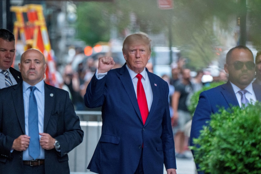 Donald Trump raises his right fist wearing a suit and red tie surrounded by security