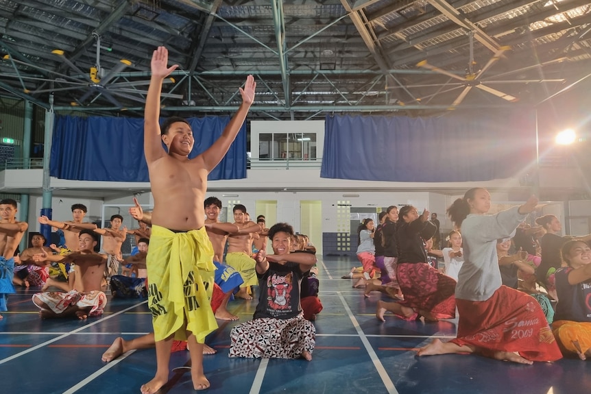 Young boy wears ie lavalava, hands outstretched in air. Students sit around him in school hall, blue court floors, behind him. 