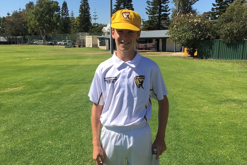 A young cricketer smiles wearing cricket whites, a yellow hat and a cricket bat.
