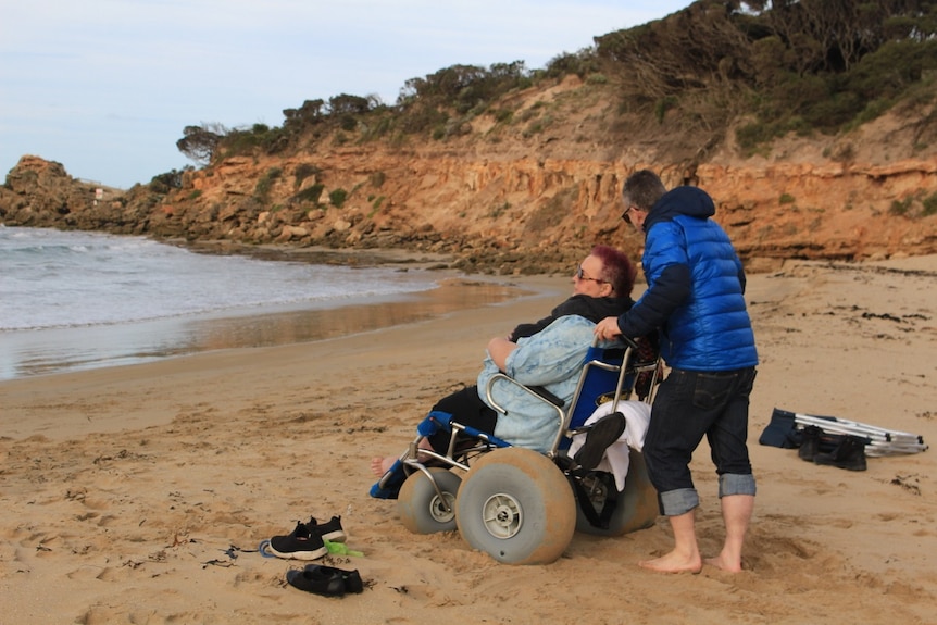 a woman in a wheelchair at a beach with someone standing behind her. 