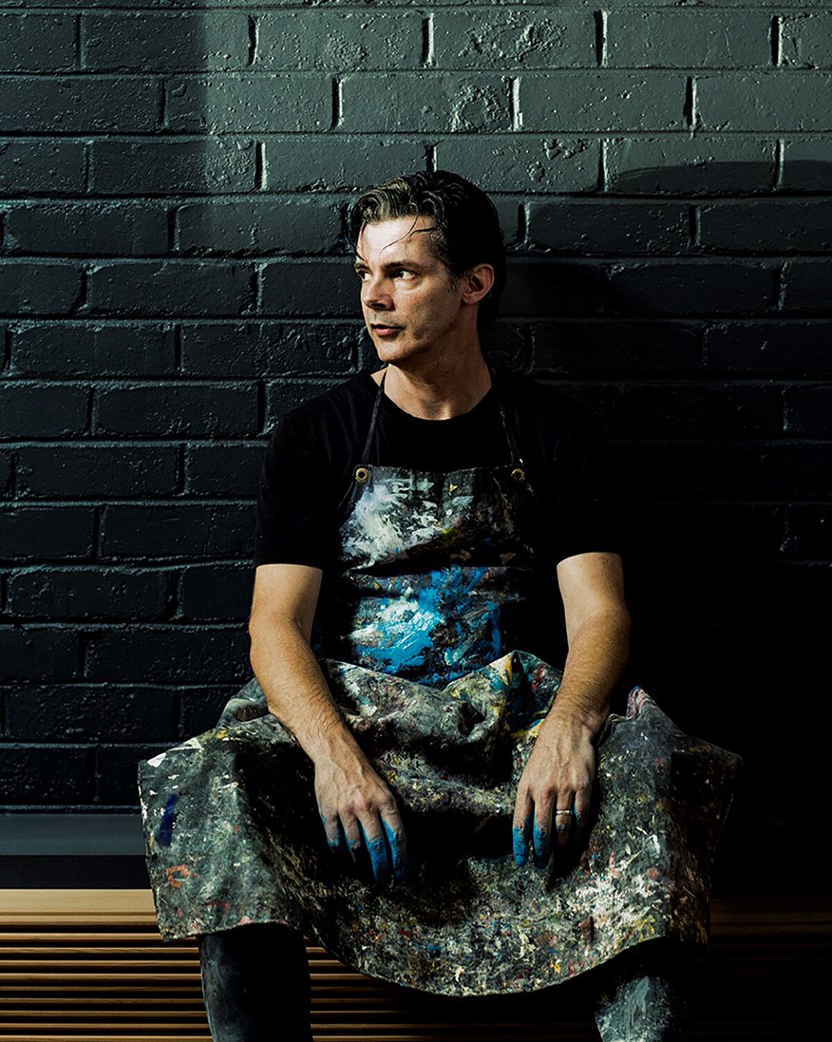 Colour photograph of Paint-maker and author David Coles wearing a paint smattered apron and sitting against a black wall.
