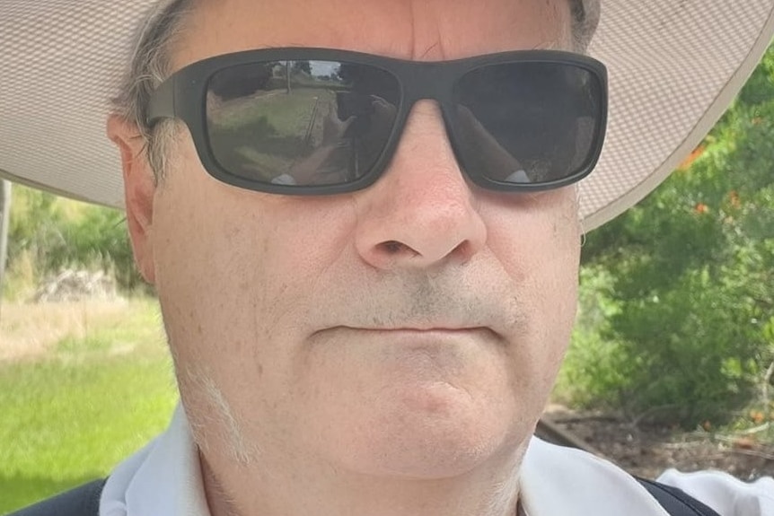 A man wearing a broad-brimmed hat and dark glasses takes a selfie outside.