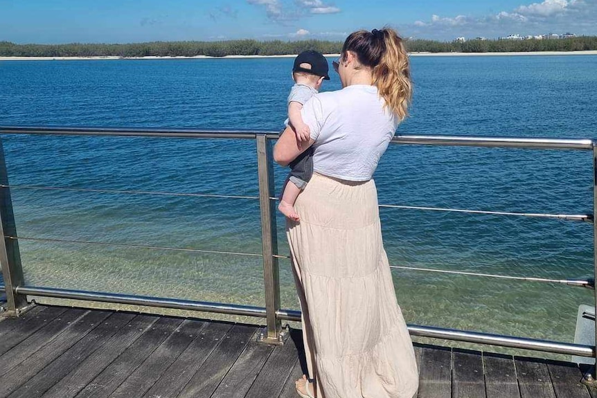 A woman holds a toddler on her hip while looking out to sea with her back to the camera.