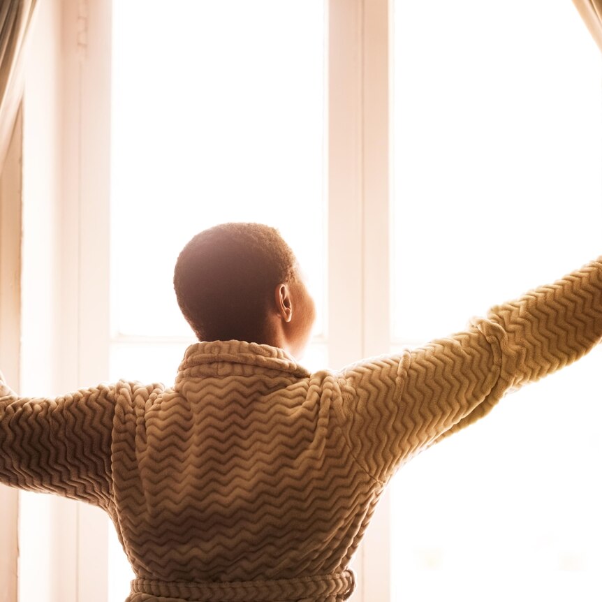 view of a woman from behind opening curtains to morning sun