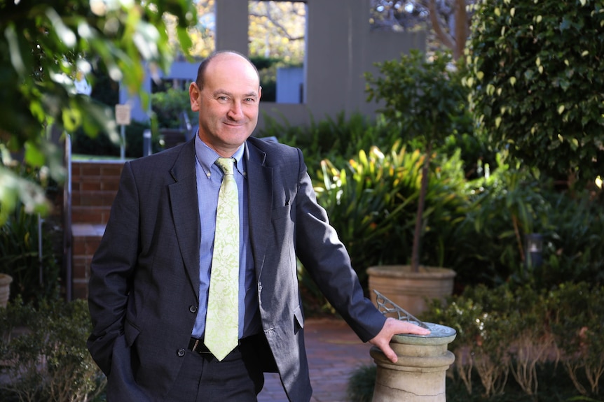 A smiling middle aged man wearing a suit and yellow tie leans up against a post outside a home.