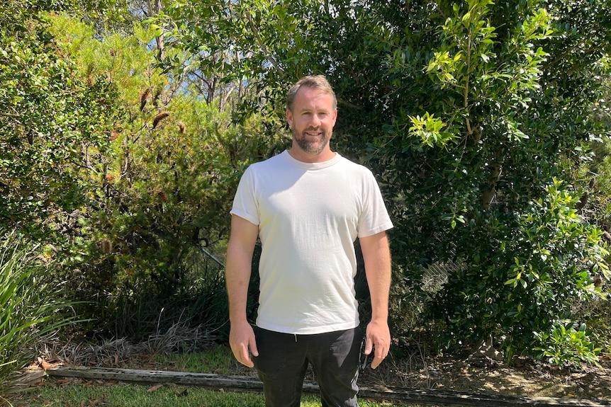 A bearded man with greying hair stands in a garden, smiling.