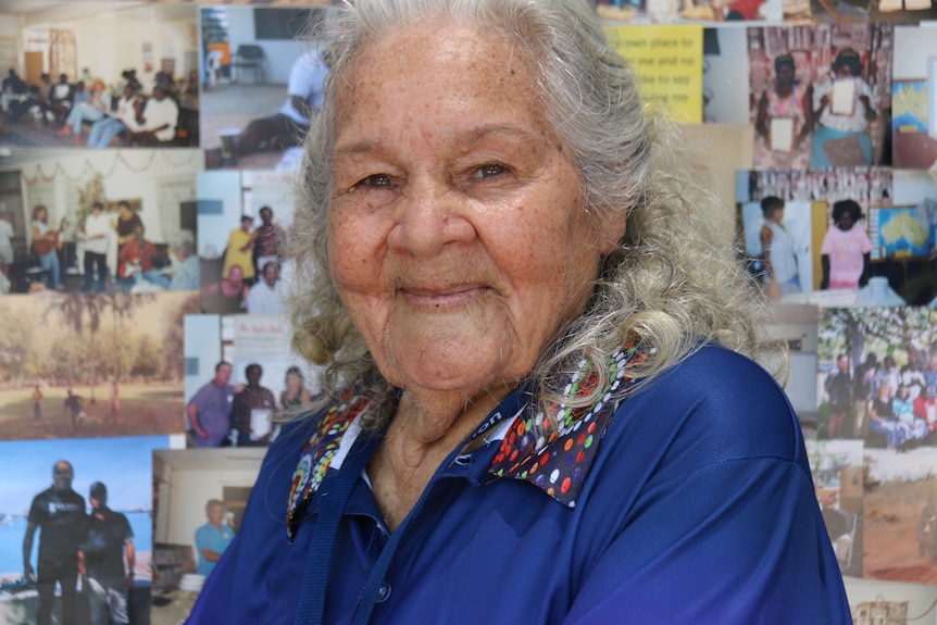 An elderly woman looks at the camera with a wall full of old photographs behind her.