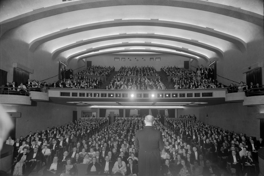 A black and white photo of a packed theatre and a man on stage facing the audience.