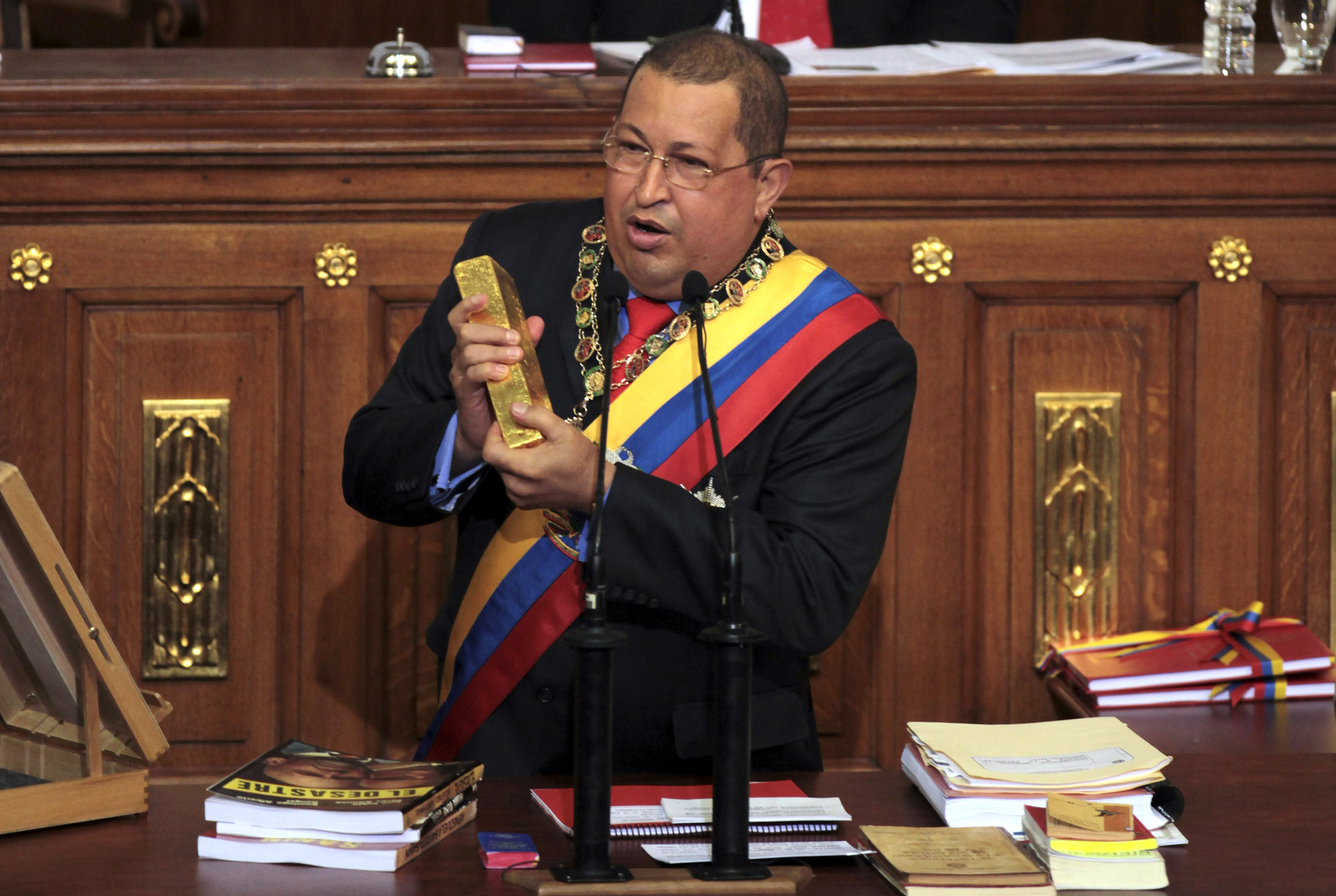 Hugo Chavez in a black suit, wearing a Venezuelan flag sash and golden necklace, holding a gold bar and speaking.