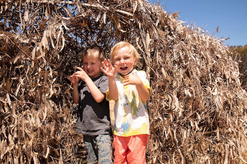 Two children pull faces from inside a dried leaf and twig hut.