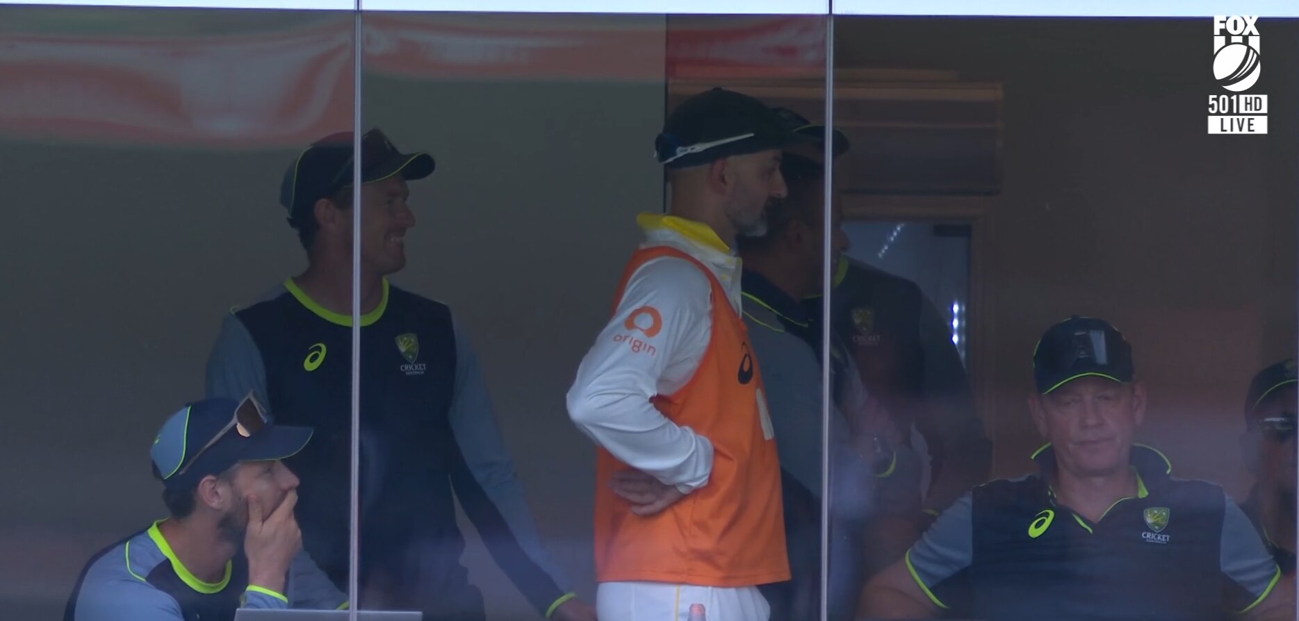 Un hombre vestido con una llamativa camiseta naranja sobre un equipo de críquet blanco se encuentra en un vestuario con las manos en las caderas.