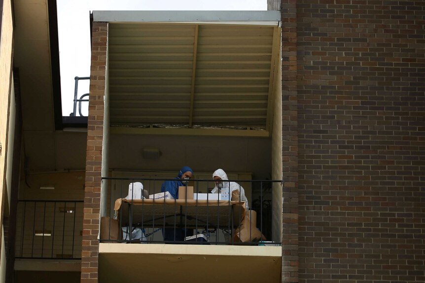 Police in overalls sit at a table on the balcony an upper floor of an apartment block.