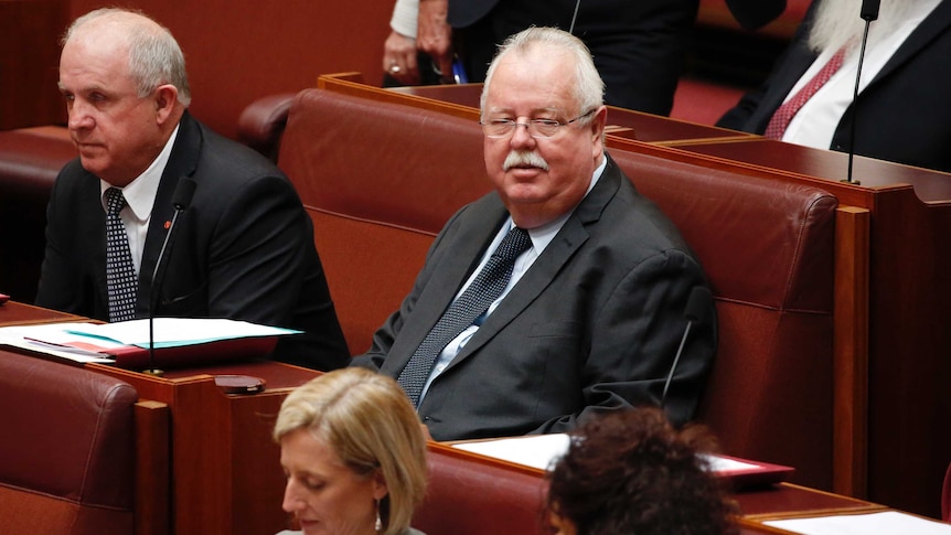 O'Sullivan has his head up, sitting next to Senator John Williams. He's wearing a black suit and glasses.