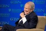 A man sitting on a couch with a blue backdrop with the initials CBN. 