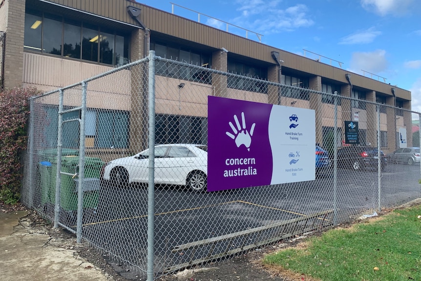A purple Concern Australia sign hangs on a chain fence in front of a car park and building.