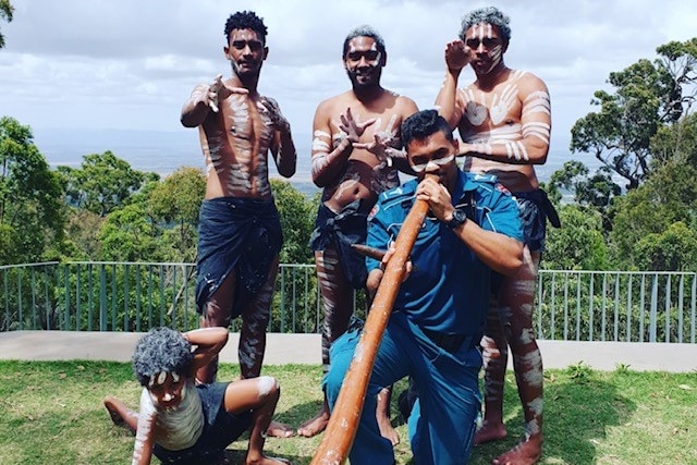 A man in paramedic clothing plays a didgeridoo with three Indigenous men in traditional paint behind him