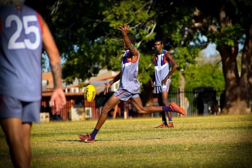 A First Nations man wearing a blue Australian rules football unfiform kicks a football during a game