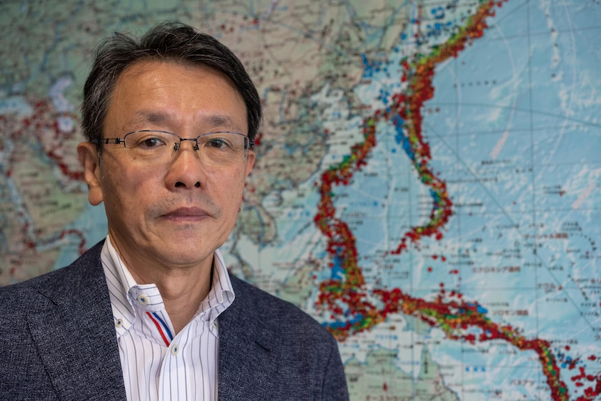 A man in wire-rimmed glasses and blazer stands in front of a map with red and orange dots all over it