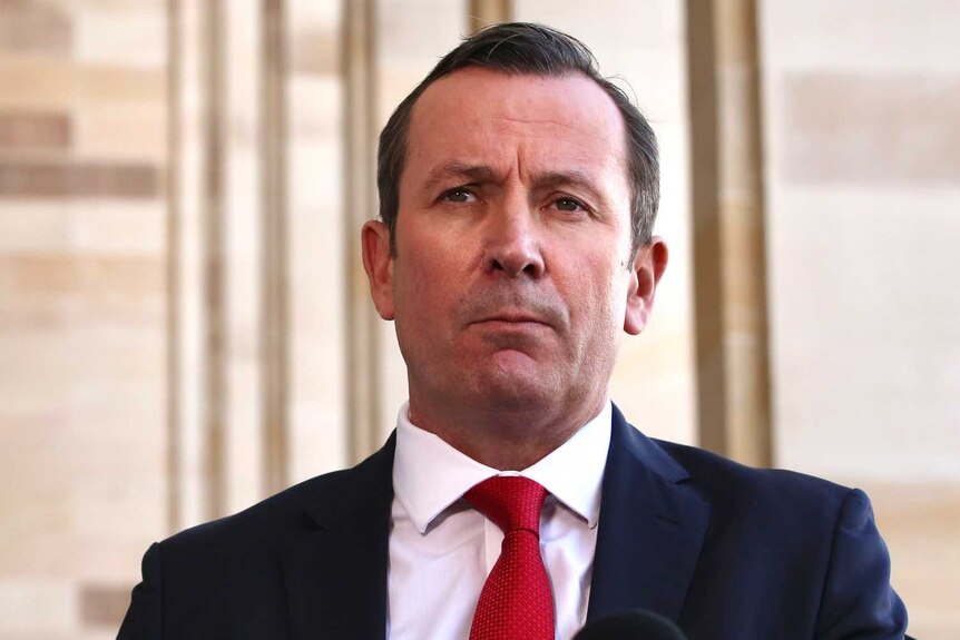 WA Premier Mark McGowan stands at a podium before microphones outside parliament wearing a red tie.