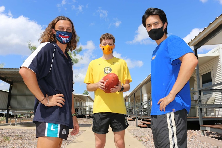 Three boys standing inside a quarantine facility holding an AFL ball outside in the sun.
